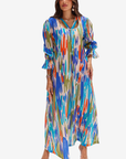 A woman modeling a long-sleeve maxi dress with a vibrant multicolor ikat pattern in shades of blue, orange, green, and white, set against a plain light gray background.