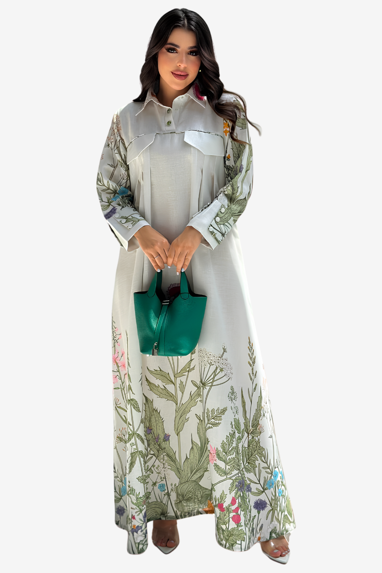A woman modeling a long-sleeved, high-neck maxi dress with a white base and green botanical leaf patterns, holding a small green handbag against a solid light gray background.