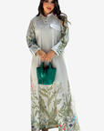 A woman modeling a long-sleeved, high-neck maxi dress with a white base and green botanical leaf patterns, holding a small green handbag against a solid light gray background.