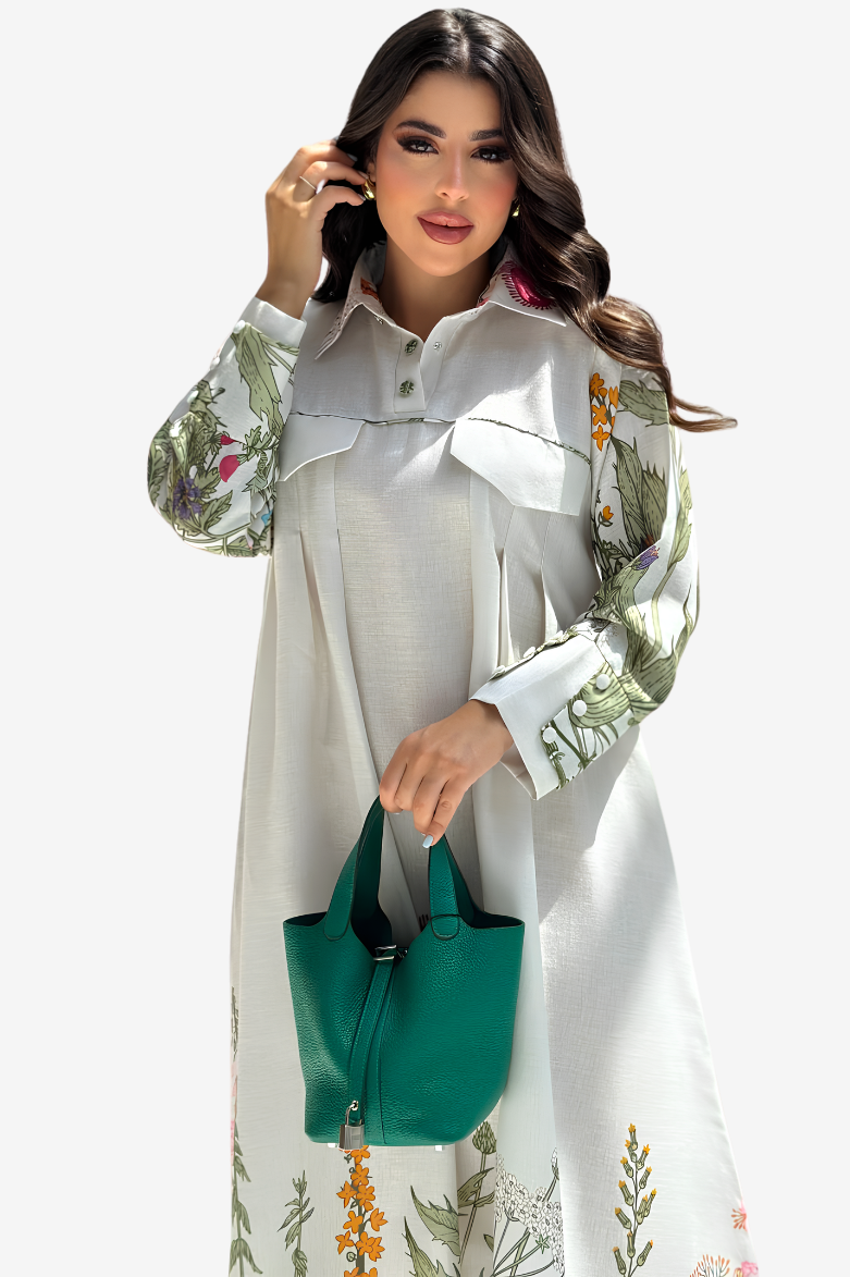 A woman modeling a long-sleeve white linen shirt dress with green botanical prints on the sleeves and hem, holding a green leather handbag against a light gray background.
