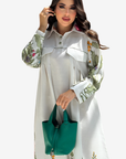 A woman modeling a long-sleeve white linen shirt dress with green botanical prints on the sleeves and hem, holding a green leather handbag against a light gray background.