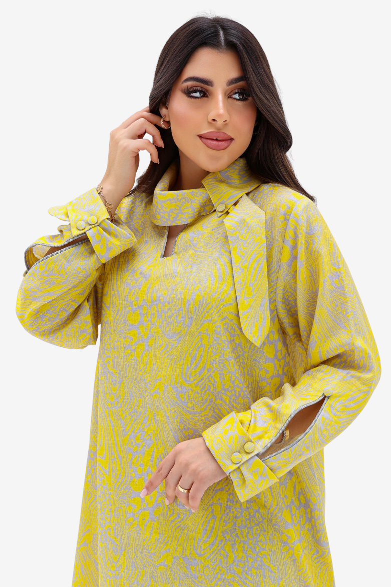 A woman modeling a yellow and gray patterned long-sleeve tunic dress with a high collar, bow detail, and button cuffs against a light gray studio background.