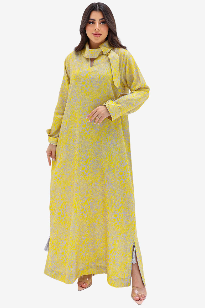 A woman modeling a long-sleeved yellow maxi dress with a subtle gray floral pattern, a tie-neck collar, and side slits, set against a plain light gray background.