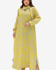 A woman modeling a long-sleeved yellow maxi dress with a subtle gray floral pattern, a tie-neck collar, and side slits, set against a plain light gray background.