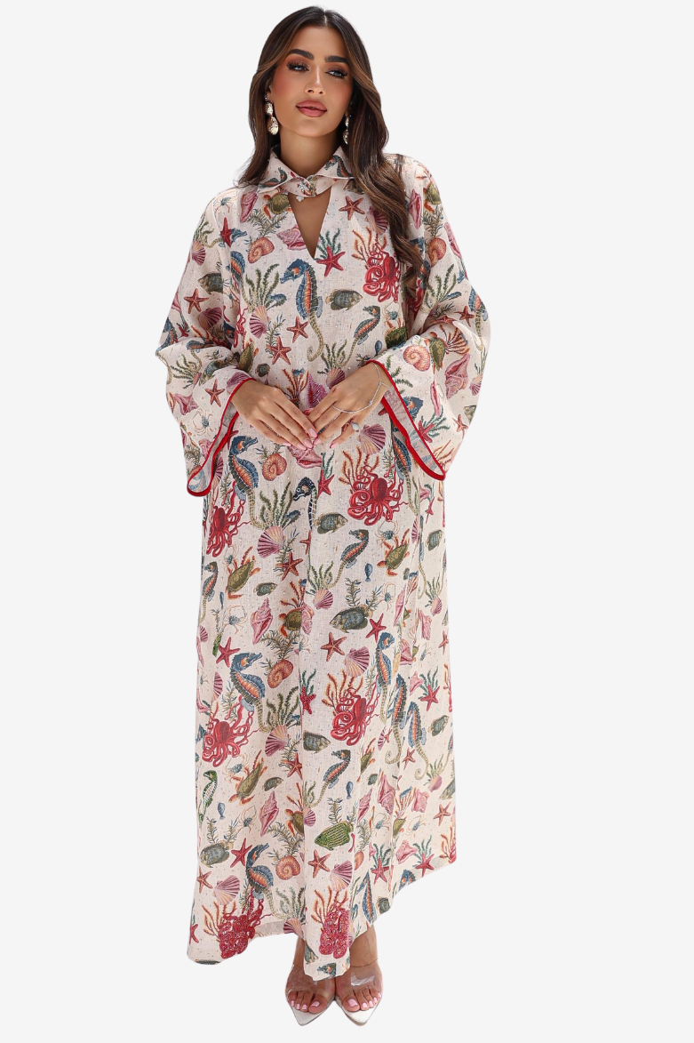 A woman modeling a long-sleeved maxi dress with a colorful coral and sea life print on a cream background, featuring red piping along the sleeve edges and a keyhole cutout at the collar.