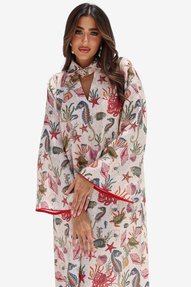 A woman modeling a long-sleeve, knee-length kaftan-style nightgown with a colorful underwater print on a cream background, featuring red piping along the cuffs and neckline.