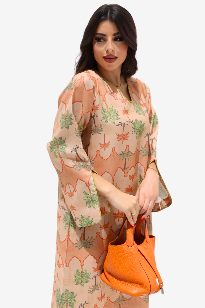 A woman modeling a peach-colored kaftan dress with a green and orange palm tree pattern, holding a bright orange handbag against a light gray background.