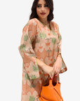 A woman modeling a peach-colored kaftan dress with a green and orange palm tree pattern, holding a bright orange handbag against a light gray background.
