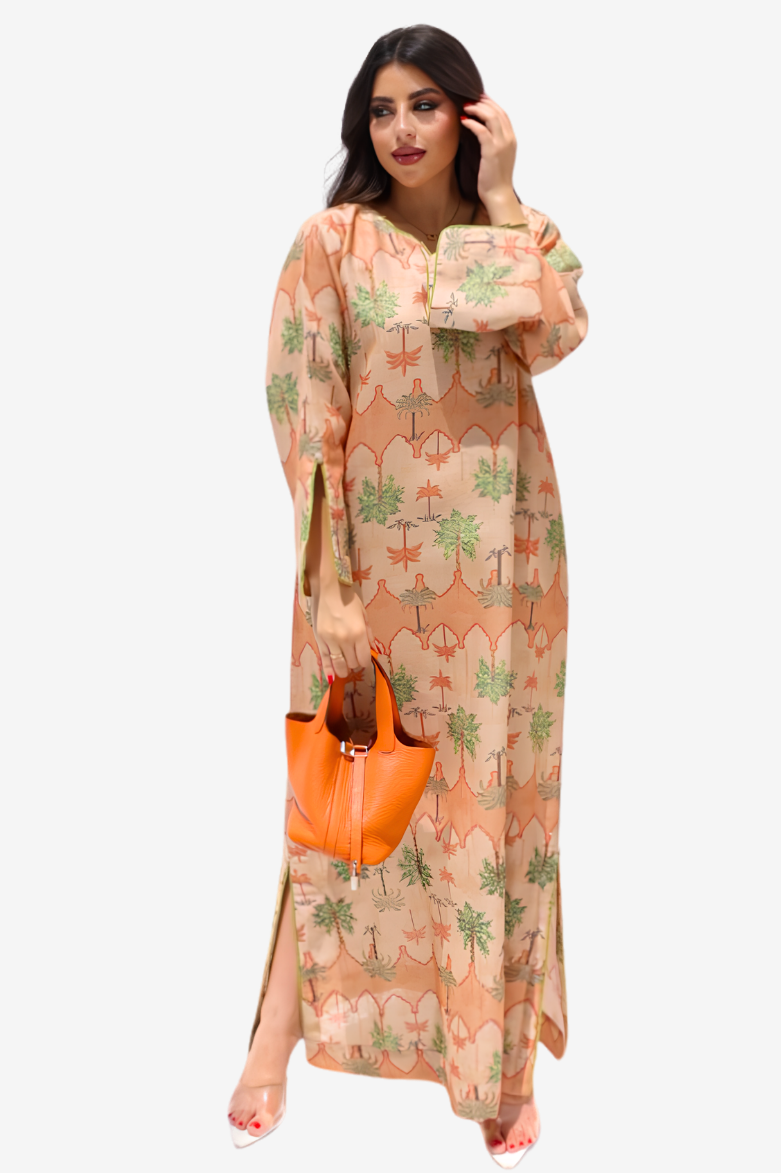 A woman modeling a long-sleeved maxi dress with a beige and orange patterned print featuring green palm tree motifs, holding an orange handbag against a light gray background.