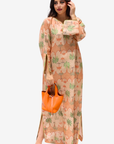 A woman modeling a long-sleeved maxi dress with a beige and orange patterned print featuring green palm tree motifs, holding an orange handbag against a light gray background.