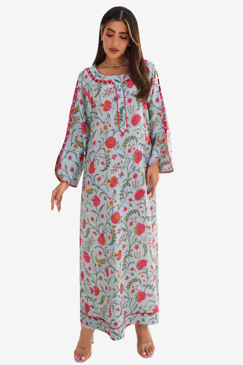 A woman modeling a long, light blue-gray kaftan dress with a vibrant pink, red, and green floral print, featuring wide bell sleeves and embroidered neckline details, set against a plain light gray background.