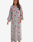 A woman modeling a long, light blue-gray kaftan dress with a vibrant pink, red, and green floral print, featuring wide bell sleeves and embroidered neckline details, set against a plain light gray background.
