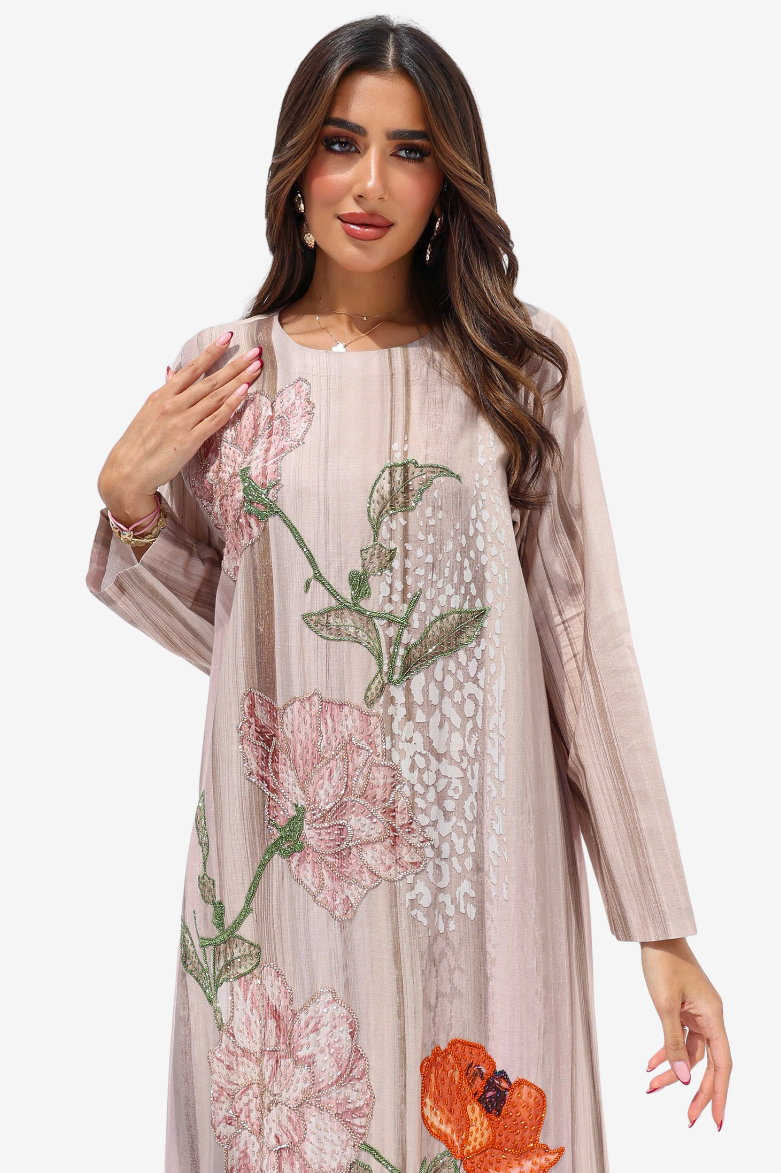 A woman modeling a long-sleeved, floor-length tunic dress in a light beige-pink color, decorated with large pink and orange floral embroidery and green leaf details on a solid white background.