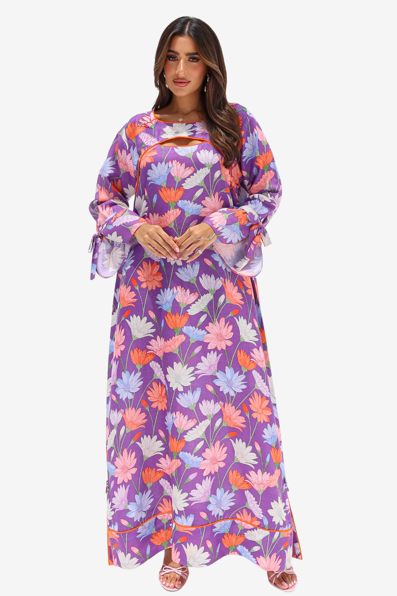 A woman modeling a long-sleeved maxi dress with a vibrant purple base and a multicolor floral print in pink, orange, and white, set against a plain light gray background.