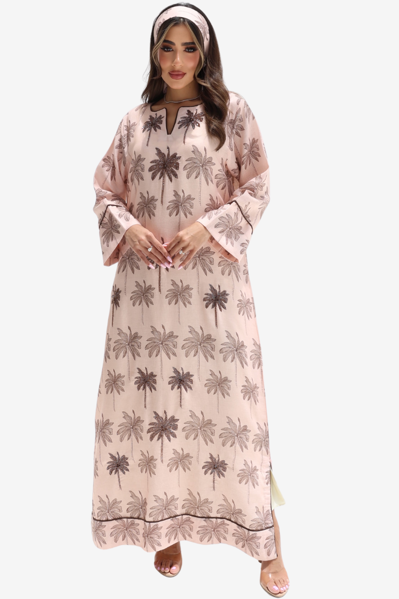 A woman modeling a long, light pink kaftan dress with a brown palm tree pattern, paired with a matching headband, against a plain light gray background.