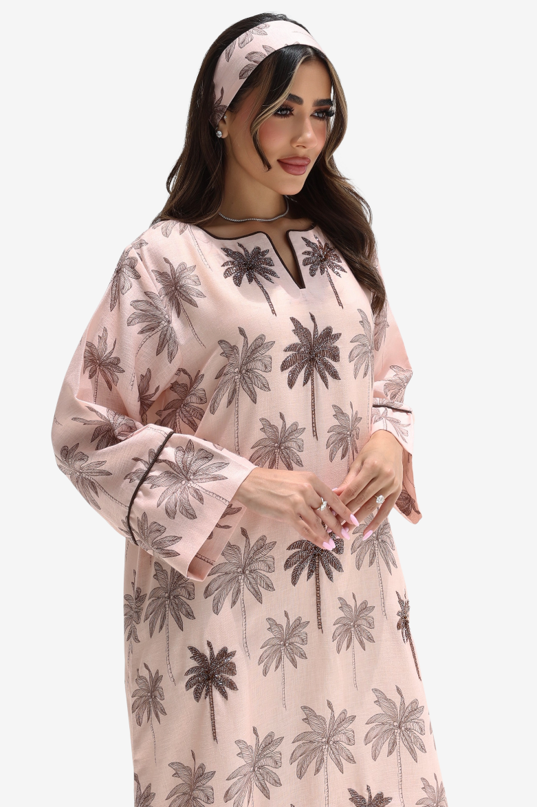 A woman wearing a light pink long-sleeve kaftan dress with a dark gray palm tree print, a matching headband, and a black choker necklace, set against a plain light gray background.