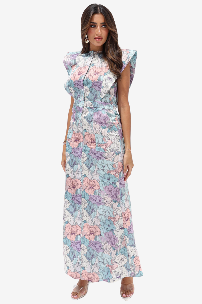 A woman modeling a sleeveless, high-neck maxi dress with a pastel floral print in pink, purple, and blue on a white background, set against a plain light gray studio backdrop.