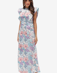A woman modeling a sleeveless, high-neck maxi dress with a pastel floral print in pink, purple, and blue on a white background, set against a plain light gray studio backdrop.