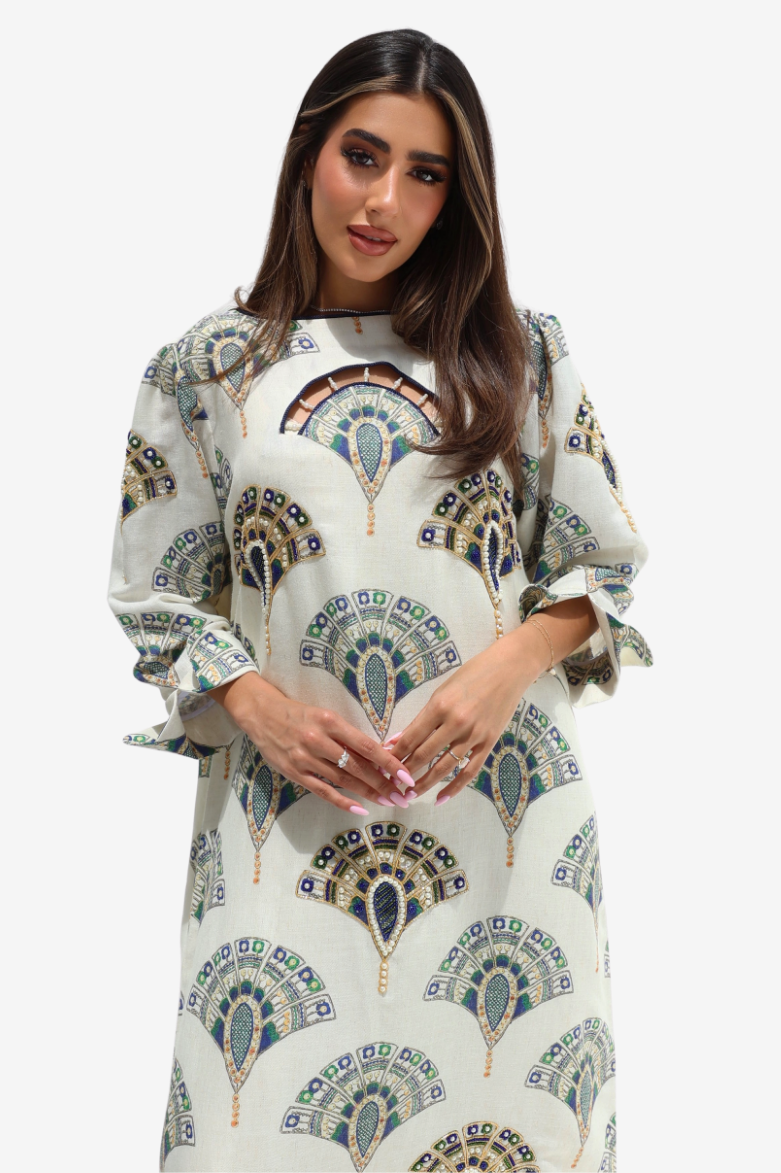 A woman modeling a long-sleeved, high-neck kaftan dress with a cream base and an intricate blue, green, and gold paisley pattern, set against a plain light gray background.
