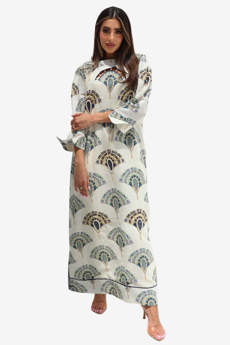 A woman modeling a long cream-colored maxi dress with a green and gold peacock feather print, featuring a high neck and flared sleeves, set against a plain light gray background.