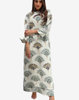 A woman modeling a long cream-colored maxi dress with a green and gold peacock feather print, featuring a high neck and flared sleeves, set against a plain light gray background.