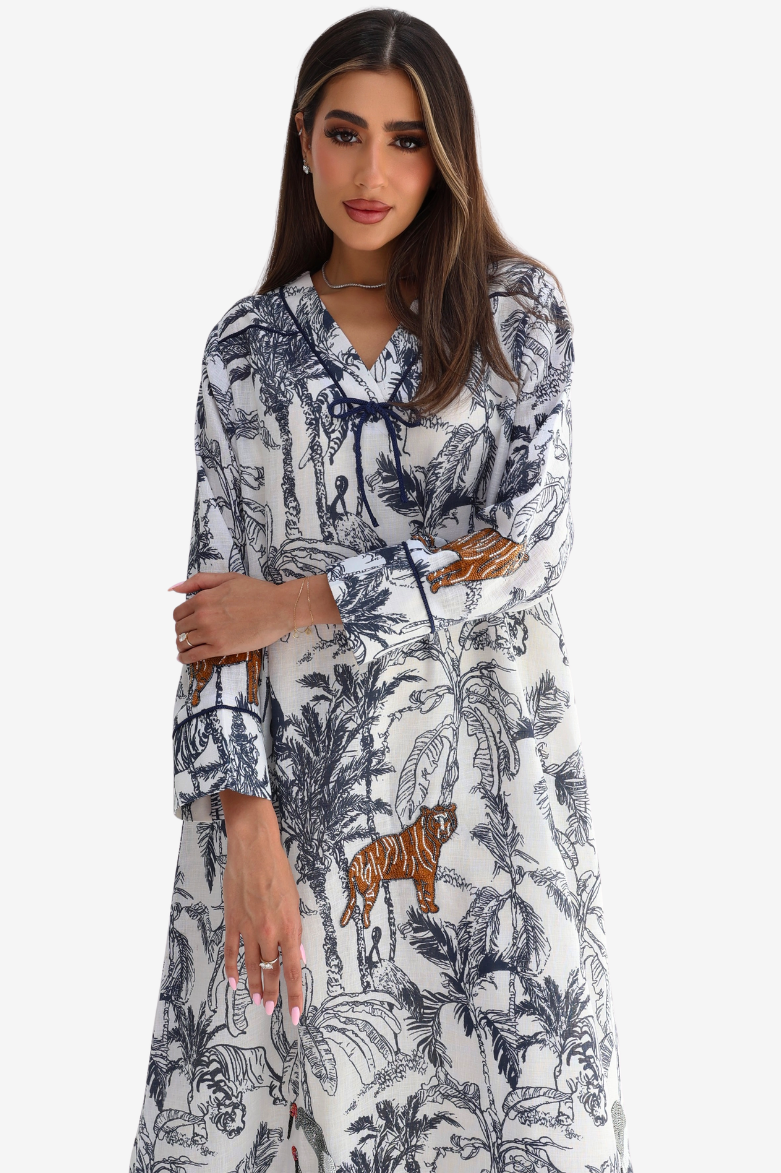 A woman modeling a long-sleeve white maxi dress with a navy blue tropical and jungle print, featuring a tiger motif near the hem.