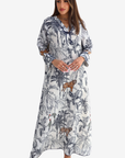 A woman modeling a long-sleeve maxi dress with a blue and brown jungle print on a white background, featuring a tie-neck detail and cutout sleeves.