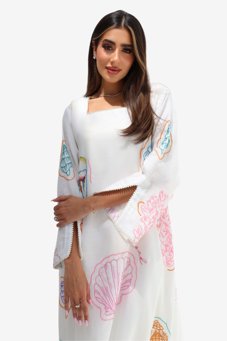 A woman modeling a white kaftan dress with colorful pastel embroidery featuring seashells, flamingos, and tropical motifs, set against a plain white background.