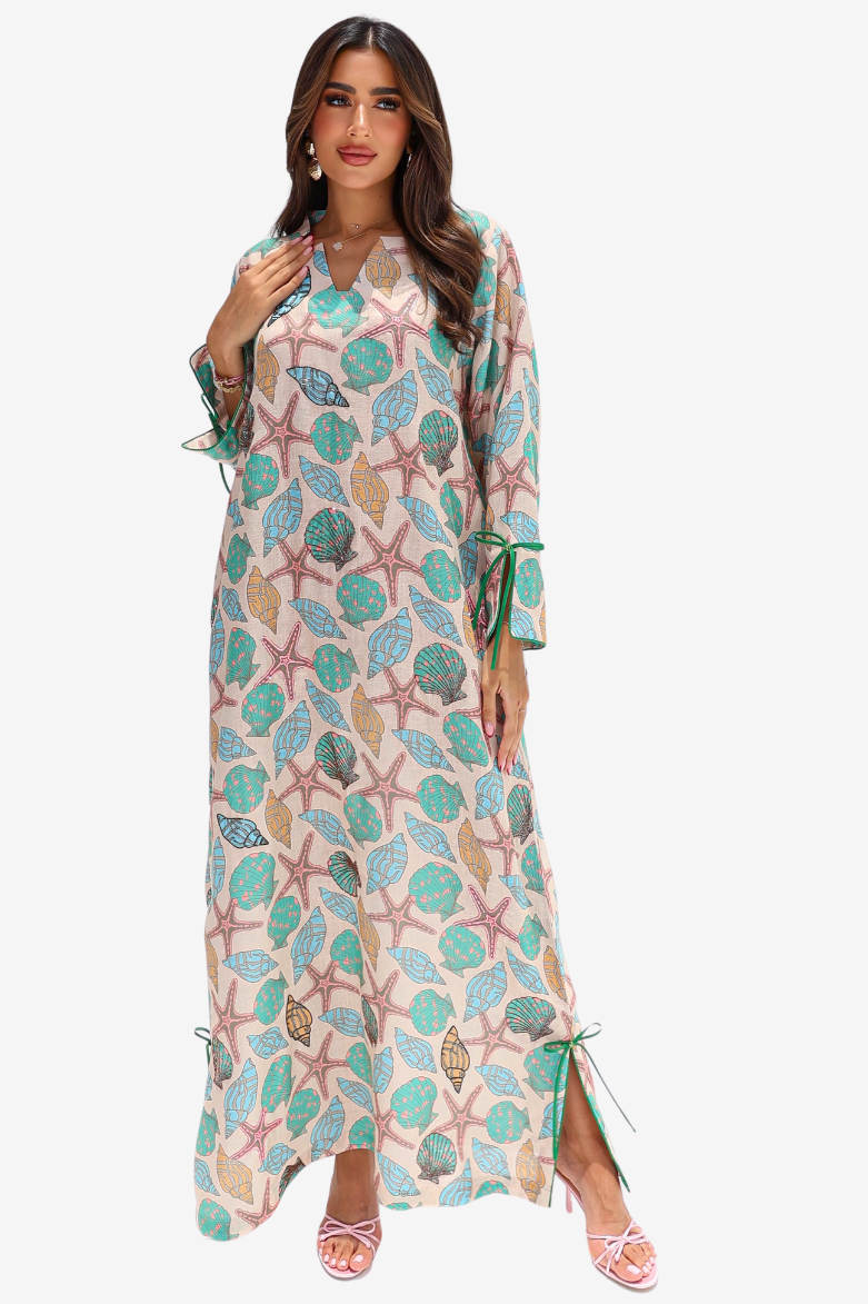 A woman modeling a long maxi dress with a seashell and starfish print in teal, pink, and beige on a light cream background, set against a plain light gray studio backdrop.