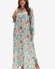 A woman modeling a long maxi dress with a seashell and starfish print in teal, pink, and beige on a light cream background, set against a plain light gray studio backdrop.