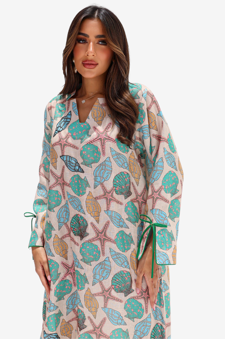 A woman modeling a long-sleeve V-neck tunic dress with a colorful seashell and starfish print on a cream background, featuring green tie details at the cuffs.